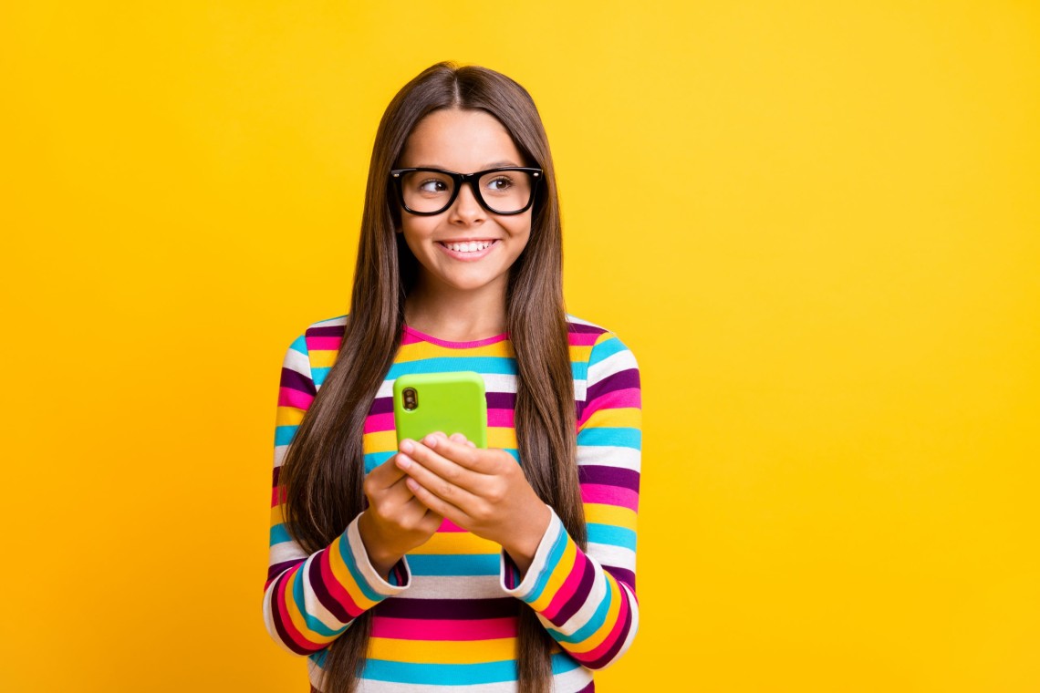 Photo portrait of curious girl keeping cellphone smiling wearing glasses looking at blank space isolated on bright yellow color background